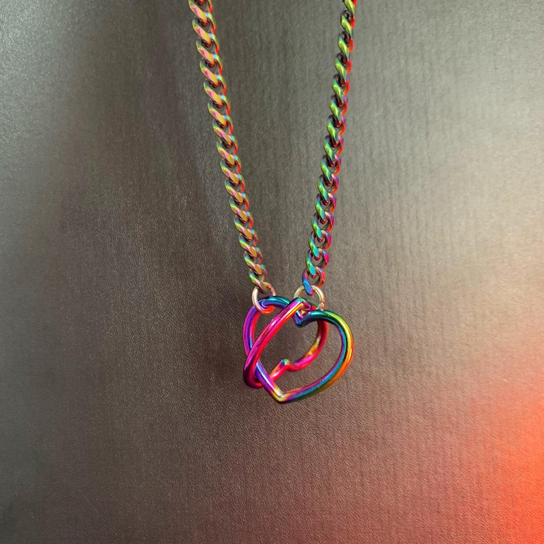 Puede incluir: Un collar que presenta una cadena de eslabones con un acabado de arco&iacute;ris iridiscente y dos colgantes entrelazados en forma de coraz&oacute;n. Los eslabones met&aacute;licos y los corazones muestran un espectro de colores azul, verde, amarillo, naranja y violeta.