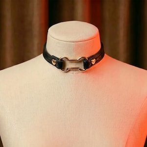 May include: Black leather choker with gold-colored hardware. The choker features a rectangular gold-colored clasp and small heart-shaped accents. The choker is displayed on a mannequin.