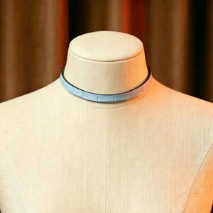 May include: A light blue choker necklace with a textured, crocodile-like pattern. The necklace is displayed on a mannequin. The choker is approximately 0.5 inches wide and has a black border.