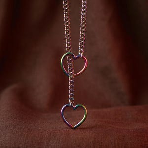 Puede incluir: Un collar con cadena de eslabones tipo barbada con un acabado iridiscente de arco&iacute;ris. La cadena pasa a trav&eacute;s de un dije en forma de coraz&oacute;n y termina en un segundo dije de coraz&oacute;n en la parte inferior. El metal muestra un espectro de colores.
