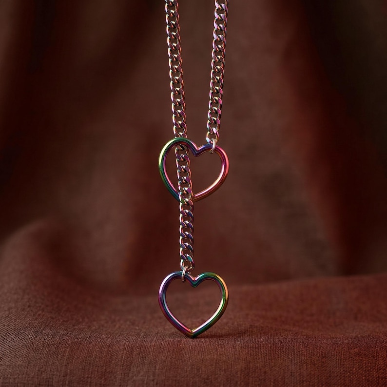 Puede incluir: Un collar con cadena de eslabones tipo barbada con un acabado iridiscente de arco&iacute;ris. La cadena pasa a trav&eacute;s de un dije en forma de coraz&oacute;n y termina en un segundo dije de coraz&oacute;n en la parte inferior. El metal muestra un espectro de colores.