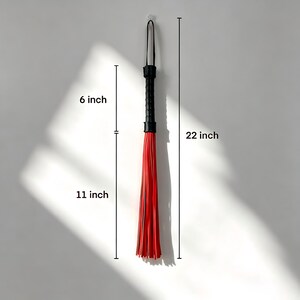May include: A black and red flogger, 56 cm long, hangs against a white wall. The handle is black, and the fall is composed of red strips. The handle is 15 cm long, and the fall is 28 cm long.