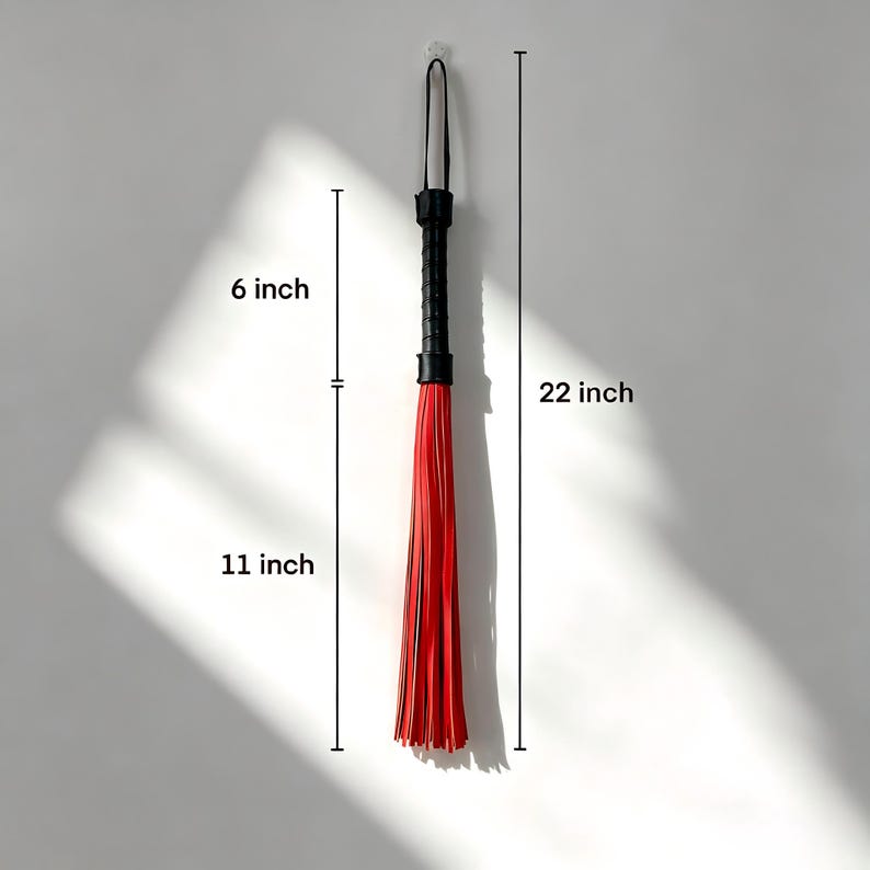 May include: A black and red flogger, 56 cm long, hangs against a white wall. The handle is black, and the fall is composed of red strips. The handle is 15 cm long, and the fall is 28 cm long.