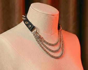 Può includere: Collana girocollo in pelle nera con borchie in metallo color argento e tre catene a strati. La collana è esposta su un manichino. La collana ha un'estetica punk rock o alternativa.