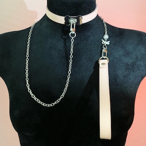 May include: A light pink collar and leash set. The collar features a bone-shaped charm and a silver chain. The leash has a matching pink handle and a decorative charm with a skull and crossbones.