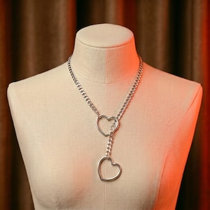 May include: Silver-toned necklace featuring two heart-shaped pendants. The necklace has a curb chain design and is displayed on a mannequin. The hearts are connected by a shorter chain, creating a layered effect. The jewelry is simple and elegant.