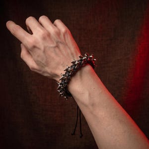 May include: A bracelet featuring a series of metallic, silver-toned, thorn-like or spiked elements arranged along a black cord. The design includes two thin, dangling black cord ends extending from the main structure of the adjustable wrist accessory.