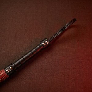 May include: A black leather whip with a handle wrapped in black leather and secured with silver rivets. The whip's end is a bundle of red leather strips. The background is a dark brown textured surface.