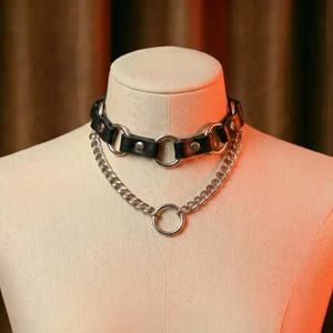 May include: A black leather choker necklace featuring silver-toned metal hardware, including circular rings and studs, layered with a separate silver-toned metal chain necklace that hangs below the choker and features a central circular ring.