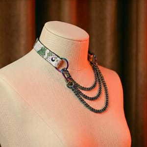 Puede incluir: Un collar tipo gargantilla blanco con un estampado de piel de serpiente en verde y gris. El collar presenta tres hilos de cadenas de color gris oscuro y anillos y cierres metálicos iridiscentes. El collar se exhibe en un maniquí.