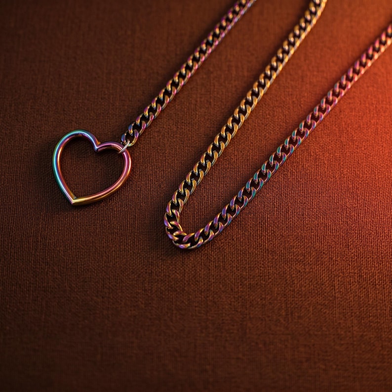 Puede incluir: Un collar con una cadena de eslabones tipo barbada y un colgante en forma de coraz&oacute;n. Tanto la cadena como el colgante presentan un acabado iridiscente de colores arco&iacute;ris que cambia entre tonos azules, morados, dorados y verdes.