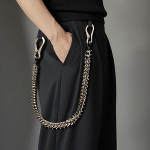 May include: A silver-toned chain belt with a spiked design hangs from black pants. The chain is attached to the pants with silver-colored clasps and black leather straps. The pants are black and have a pocket.