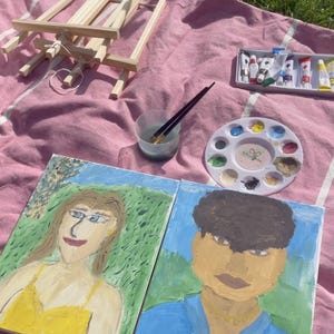 May include: An outdoor art setup on a pink blanket. Includes two painted portraits, a wooden easel, paint tubes, a palette with paint, and paintbrushes. The portraits depict a person in a yellow dress and another in a blue shirt.
