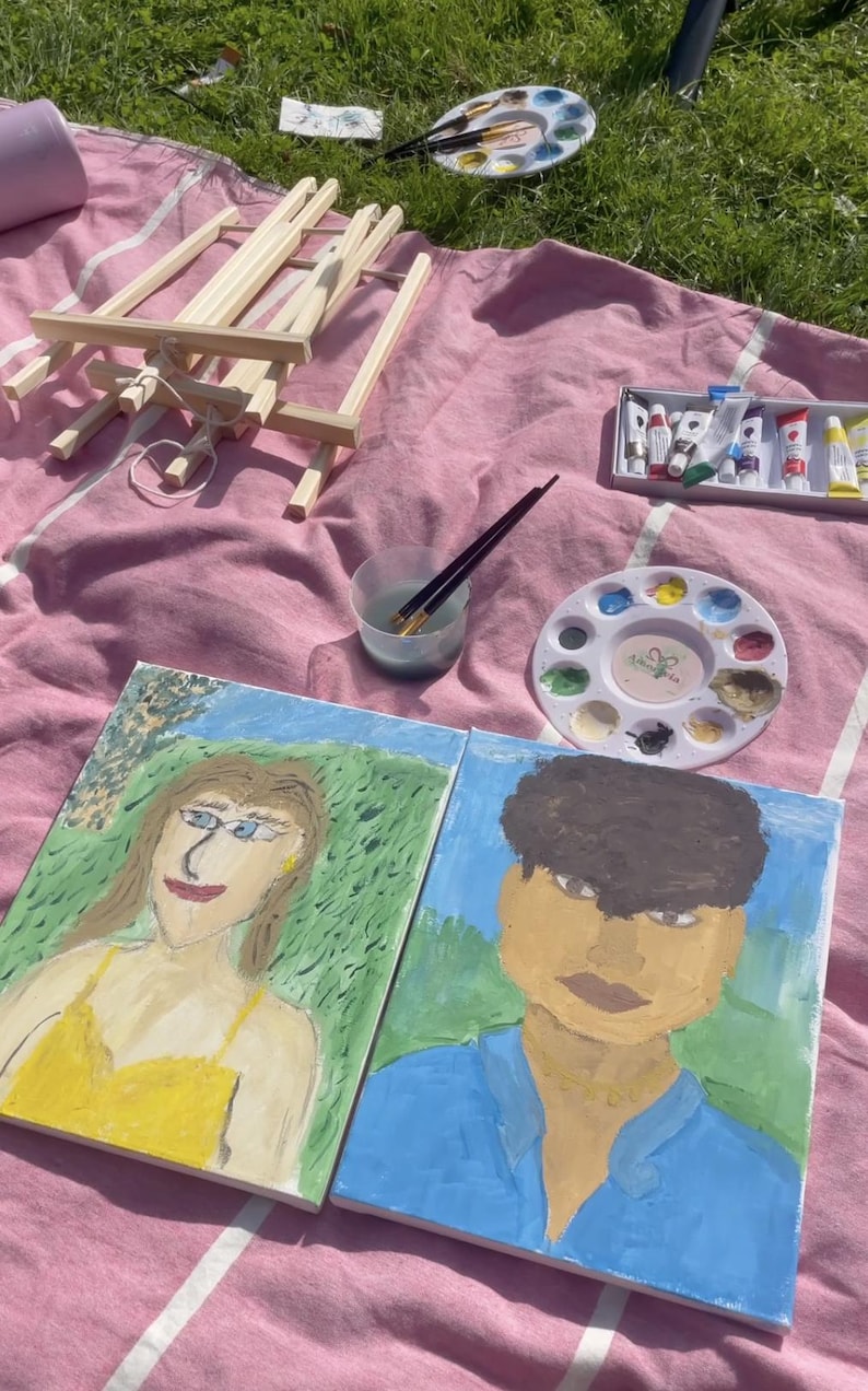 May include: An outdoor art setup on a pink blanket. Includes two painted portraits, a wooden easel, paint tubes, a palette with paint, and paintbrushes. The portraits depict a person in a yellow dress and another in a blue shirt.
