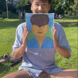 May include: A painting of a person with brown hair and a blue shirt is held up in front of a person. The painting has a blue background with a green base. The person holding the painting is wearing a light blue button-down shirt and khaki shorts.