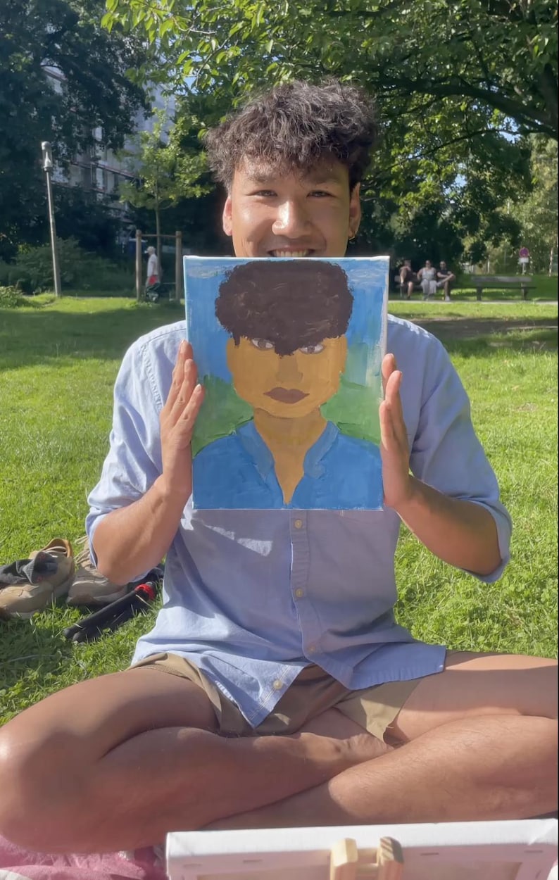 May include: A painting of a person with brown hair and a blue shirt is held up in front of a person. The painting has a blue background with a green base. The person holding the painting is wearing a light blue button-down shirt and khaki shorts.
