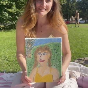 May include: A painting of a person with blonde hair and a yellow dress, set against a green and blue background. The painting is held by a person with long, auburn hair, sitting on a pink blanket outdoors.