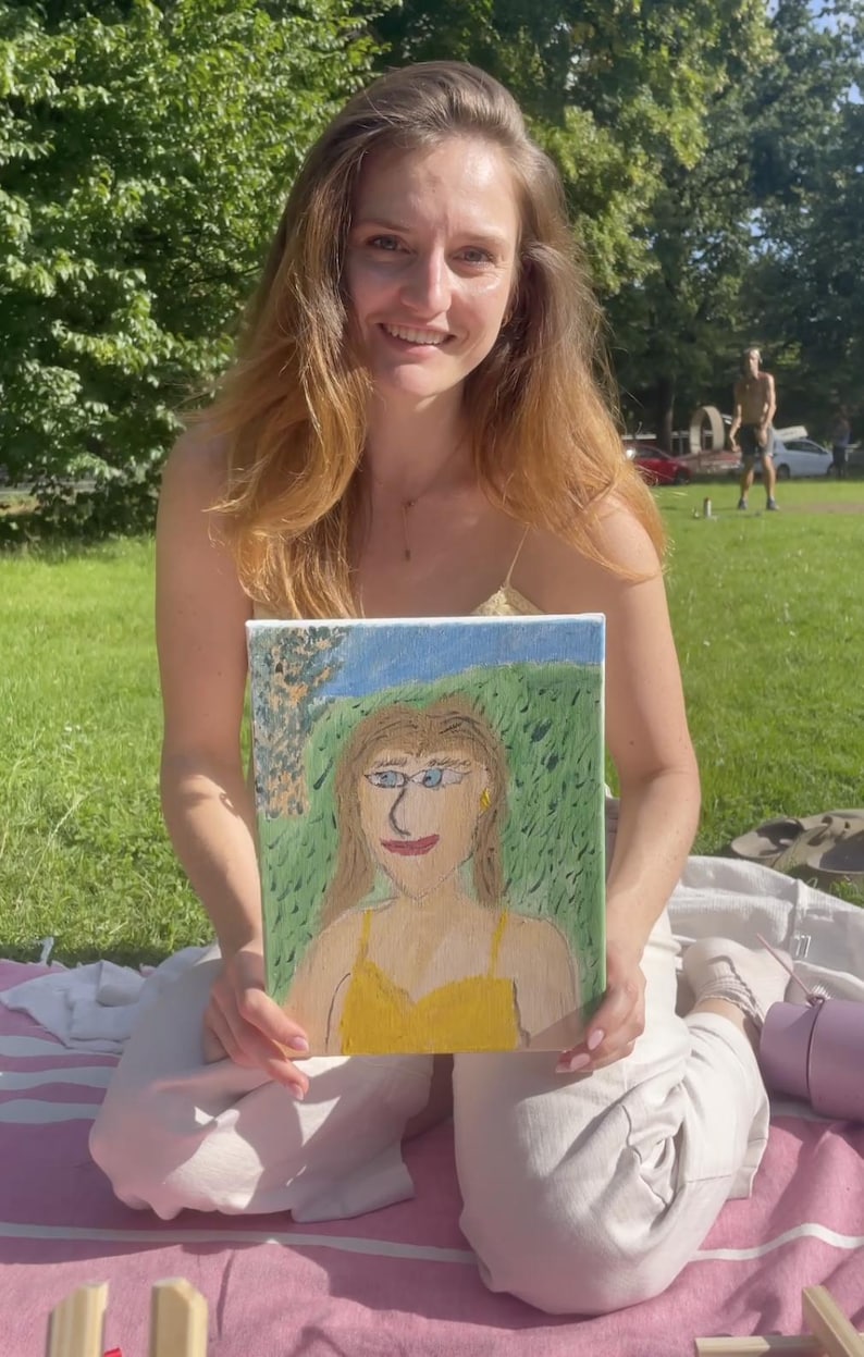 May include: A painting of a person with blonde hair and a yellow dress, set against a green and blue background. The painting is held by a person with long, auburn hair, sitting on a pink blanket outdoors.