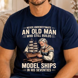 May include: A navy blue t-shirt featuring a graphic of a gray-haired person building a wooden model ship on a workbench. The text reads: NEVER UNDERESTIMATE AN OLD MAN WHO STILL BUILDS MODEL SHIPS IN HIS SEVENTIES, printed in white and light blue.