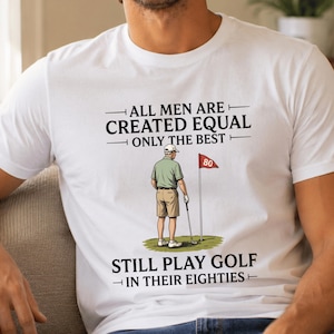 May include: A white t-shirt featuring a graphic of a golfer on a green with a flag marked 80. The text reads: ALL MEN ARE CREATED EQUAL ONLY THE BEST STILL PLAY GOLF IN THEIR EIGHTIES. The design includes black lettering and a colorful illustration.