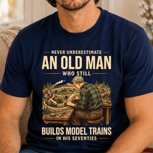 May include: A navy blue t-shirt featuring a graphic illustration of a person working on a model train set. The text reads: NEVER UNDERESTIMATE AN OLD MAN WHO STILL BUILDS MODEL TRAINS IN HIS SEVENTIES. The design includes a train, tracks, and trees.