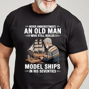 May include: A black short-sleeved t-shirt featuring a graphic of a person building a model ship. The text reads: NEVER UNDERESTIMATE AN OLD MAN WHO STILL BUILDS MODEL SHIPS IN HIS SEVENTIES. The graphic shows a person in a patterned sweater.
