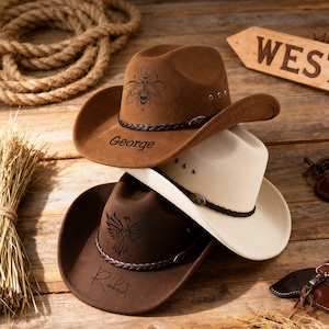 May include: Three cowboy hats stacked on a wooden surface. The top hat is brown with a bee design and the name "George". The middle hat is cream-colored. The bottom hat is brown with an eagle design and the name "Robert".