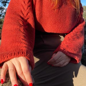 Può includere: Un maglione lavorato a maglia color ruggine con dettagli a treccia sulle maniche. Il maglione ha un girocollo e maniche lunghe. La persona indossa pantaloni verde oliva. Le unghie sono dipinte di rosso.