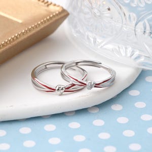 Puede incluir: Dos anillos de plata con detalles de esmalte rojo y un pequeño detalle en forma de corazón. Un anillo presenta una pequeña gema transparente. Los anillos se exhiben sobre una superficie blanca con un fondo de lunares azules.