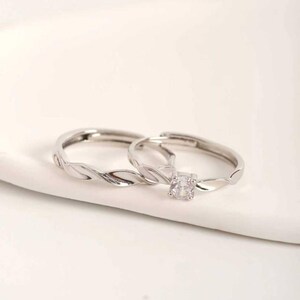 Puede incluir: Dos anillos plateados con un diseño de banda trenzada. Un anillo presenta una gema cuadrada y transparente. Los anillos se exhiben sobre una superficie blanca, mostrando su diseño elegante y sencillo.