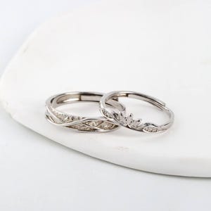 Könnte beinhalten: Zwei silberne Ringe mit aufwendigen Designs. Ein Ring hat ein geflochtenes Muster, der andere ein blattartiges Design mit kleinen, funkelnden Akzenten. Die Ringe werden auf einer weißen, strukturierten Oberfläche präsentiert.