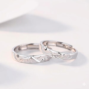 Könnte beinhalten: Zwei silberne Ringe mit eingravierten Designs. Einer zeigt Sonne und Berge, der andere Mond und Wellen. Die Ringe sind vor einem weißen Hintergrund platziert.