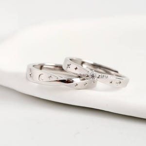Pode incluir: Dois anéis de prata com desenhos celestes. Um anel apresenta uma lua crescente e estrelas, enquanto o outro tem uma fileira de pequenas estrelas e uma pedra central em forma de diamante. Os anéis são exibidos em uma superfície branca.