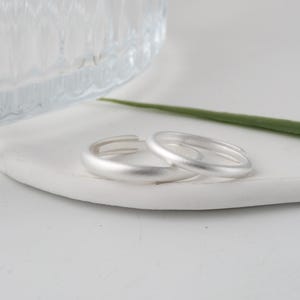 Puede incluir: Dos anillos de plata con acabado mate sobre una superficie blanca y curvada. Un recipiente de vidrio transparente y una hoja verde en el fondo. Los anillos son sencillos y elegantes.
