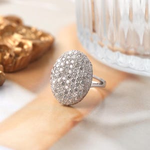 Puede incluir: Un anillo plateado con una gran cara ovalada cubierta de pequeñas piedras brillantes. El anillo se presenta sobre un fondo suave y neutro, con un objeto de vidrio borroso y un elemento decorativo dorado en el fondo.