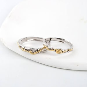 May include: Two silver rings with gold accents. The rings have a textured, organic design, resembling intertwined branches. One ring features a small, gold-colored detail. The rings are displayed on a white surface.