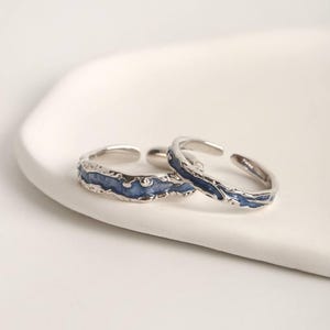 May include: Two silver-colored adjustable rings with a textured blue enamel design. The rings are displayed on a white surface. The blue enamel has a wavy, organic pattern, adding a unique touch to the jewelry.