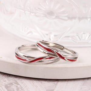 Può includere: Due anelli d'argento con un design a strisce di smalto rosso. Gli anelli sono esposti su una superficie bianca, rotonda e strutturata. Gli anelli sono probabilmente per coppie o come simbolo di impegno.