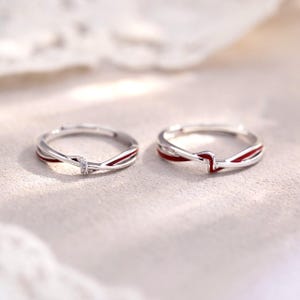 May include: Two silver-toned rings featuring a delicate twisted band design with a thin red enamel inlay. The left ring includes a small, clear, sparkling stone set at the center of the twist, while the right ring features a plain metal twist.