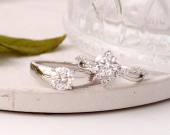 May include: Two silver rings with large, clear gemstones. One ring has a single round stone, while the other features a cluster of smaller stones around a larger, central gem. The rings are displayed on a white, circular surface.