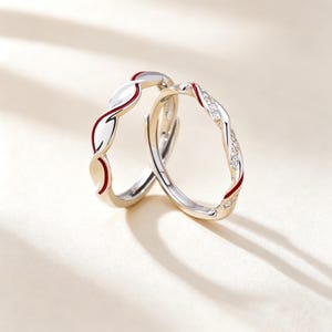 May include: Two silver-toned rings with a twisted design. One ring features a smooth metallic surface with a thin red enamel inlay, while the other ring features a twisted band set with small clear stones and a thin red enamel accent line.