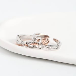 Puede incluir: Un conjunto de dos anillos con un diseño de enredadera en tono plateado con hojas pequeñas y un detalle de flor de rosa en tono oro rosa. Los anillos tienen una estructura de banda abierta e incluyen pequeñas piedras claras incrustadas.
