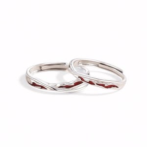 Peut inclure: Deux bagues argentées dotées d'un design ouvert et réglable. Chaque bague présente une finition métallique polie avec un canal ondulé en creux, rempli d'une incrustation en émail rouge profond qui suit le contour organique et fluide de l'anneau.