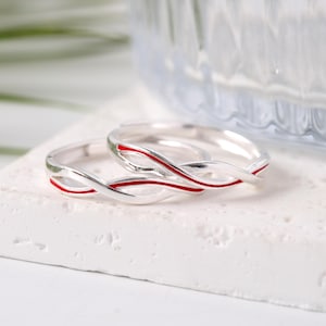 Puede incluir: Dos anillos de plata con incrustaciones de esmalte rojo. Los anillos tienen un diseño entrelazado, descansando sobre una superficie blanca. Los anillos son probablemente para una pareja, simbolizando el amor y el compromiso.