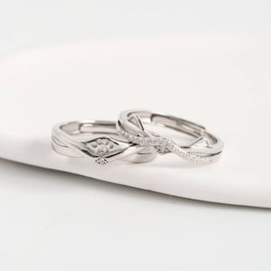 Op de afbeelding: Twee zilveren ringen met een pootafdrukontwerp en kleine heldere stenen. Eén ring heeft een pootafdruk en de andere heeft een golfontwerp met kleine stenen. De ringen liggen op een wit oppervlak.