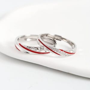 Può includere: Due anelli d'argento con un design a strisce di smalto rosso. Un anello presenta una fila di piccole pietre trasparenti. Gli anelli sono su uno sfondo bianco, mostrando il loro design elegante. Ideale per le coppie.