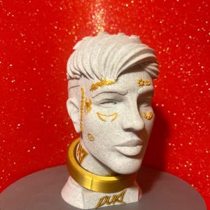 Puede incluir: Busto blanco con detalles dorados, incluyendo acentos faciales y un collar. El busto presenta cabello corto y estilizado. La base tiene la palabra "DUK" en dorado. Fondo rojo brillante.