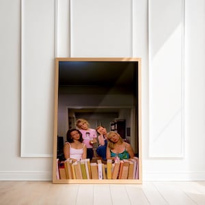 May include: A framed print featuring a group of four people leaning over a row of books. The print has a light wooden frame and is set against a white wall and wooden floor. The people are wearing various tops and dresses. The image is a photograph.