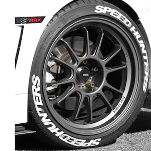 May include: Close-up of a car wheel with a dark gray rim and a black tire. The tire has white lettering that says "SPEED HUNTERS". The rim has a multi-spoke design. The car is white and has "WRX" printed on the side.