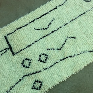 Puede incluir: Una alfombra rectangular blanquecina con una superficie texturizada y patrones geométricos negros. El diseño incluye líneas rectas y anguladas, junto con pequeñas formas florales estilizadas. La alfombra probablemente está hecha de un material suave y tejido.